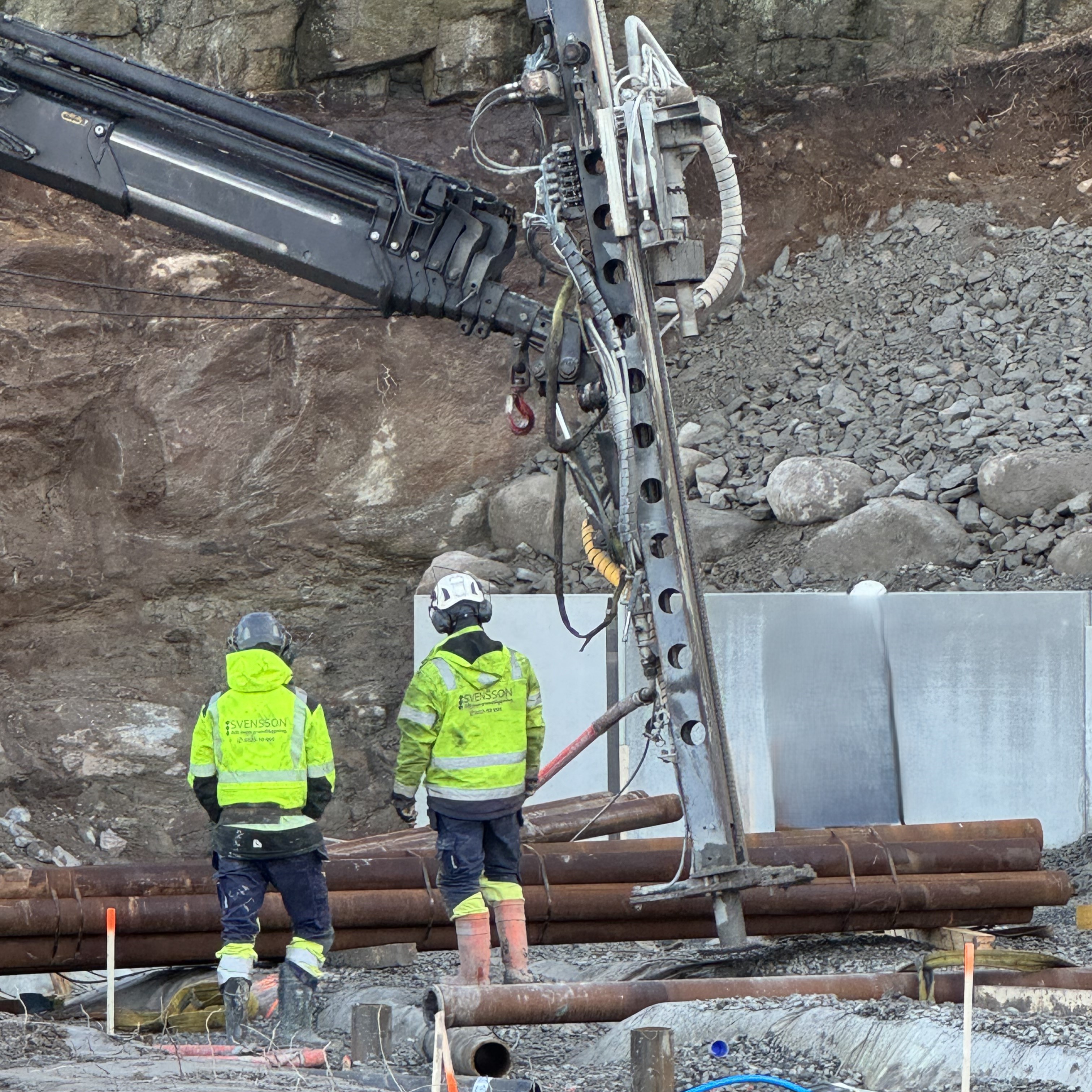 Två personer i varselkläder och hjälm samt en pålmaskin