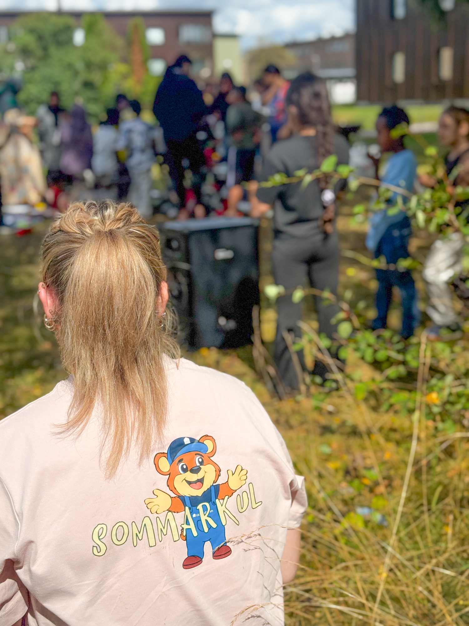 I förgrunden ser man ryggen på en av aktivitetsvärdarna på sommarkul, i bakgrunden syns fullt av människor som köar för att få sjunga
